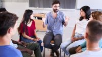 Diverse group of high school students sitting on chairs in a circle and interacting during a lesson, their Caucasian male teacher with them and talking. Education concept