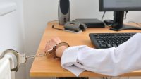 Detail of business person hand tied with handcuffs to workplace keyboard and monitor in background.