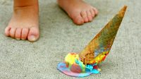 a dropped rainbow colored ice cream cone lays upside down on the sidewalk at the feet of a young child
** Note: Shallow depth of field