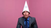 A businessman sat in the corner of the room wearing a dunce's hat with a dumb expression on his face.